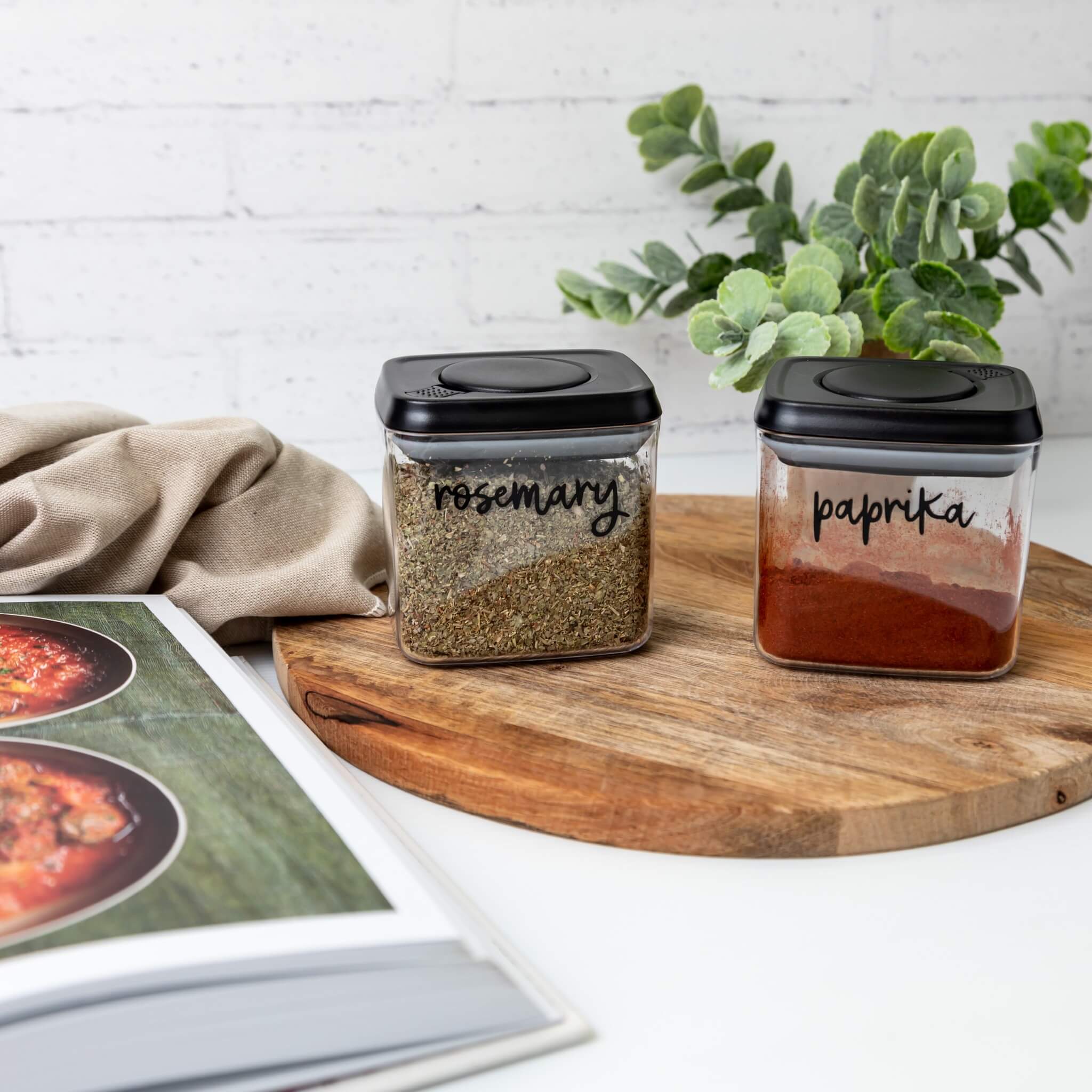 Two spice containers labeled 'rosemary' and 'paprika' on a wooden board with a magazine and plant in the background.