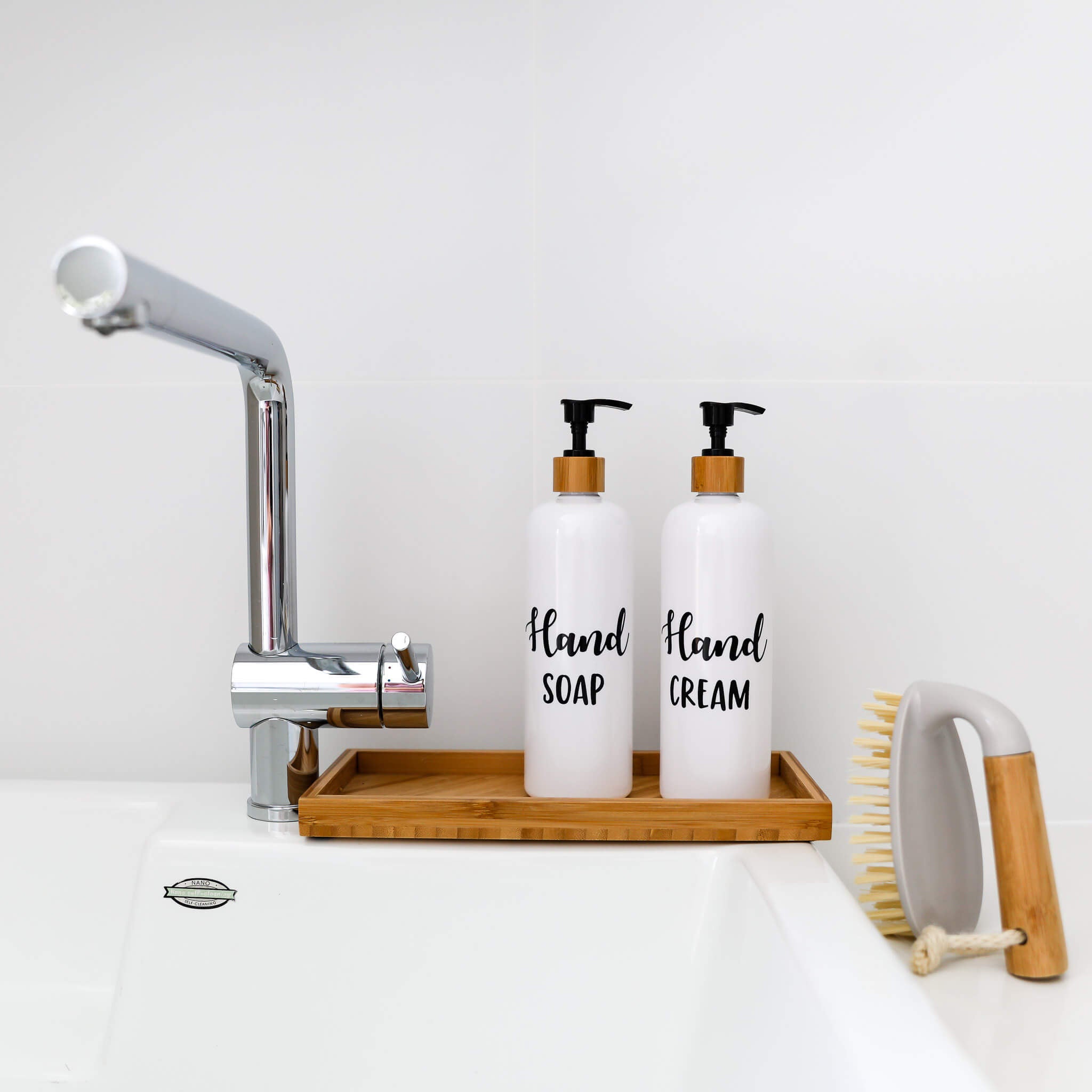 Two white pump bottles labeled 'HAND SOAP' and 'HAND CREAM' on a wooden tray next to a dish brush, with a tap in the background.