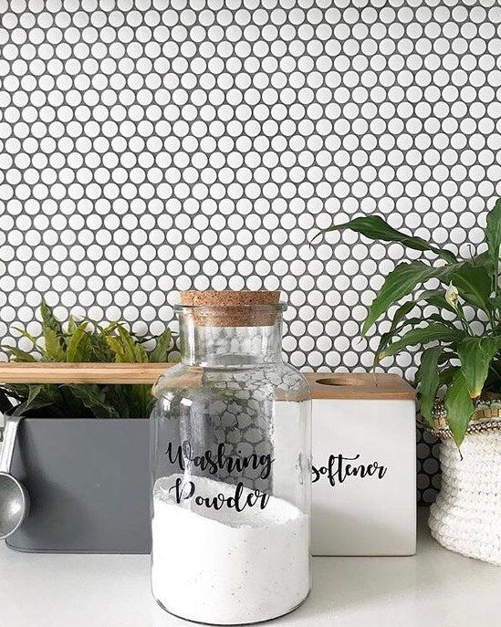 Custom vinyl laundry labels on glass jars with washing powder and softener, set against a white tiled background with green plants.