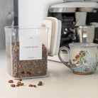 Container of coffee beans with a coffee machine and mug in the background