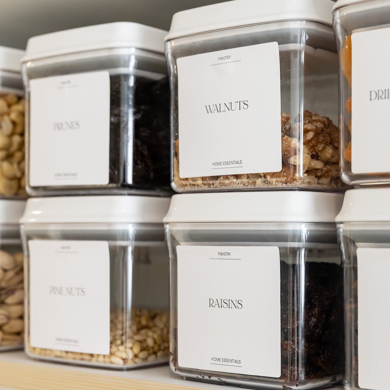 Pantry storage containers with labels on a shelf containing various nuts and seeds.