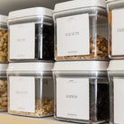 Pantry storage containers with labels on a shelf containing various nuts and seeds.