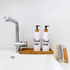 Two white pump bottles labeled 'HAND SOAP' and 'HAND CREAM' on a wooden tray next to a dish brush, with a tap in the background.