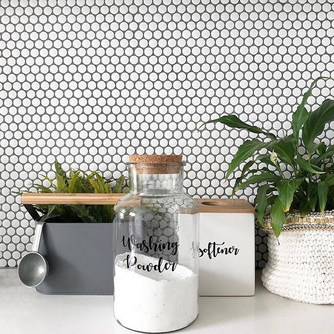 Custom vinyl laundry labels on glass jars with washing powder and softener, set against a white tiled background with green plants.